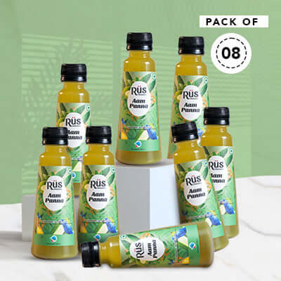 Rus Organic Aam Panna cold-pressed juice is angled on a bed of light-colored sand with scattered seashells and small stones. The bottle has a black cap and a green and light blue label featuring illustrations of green mangoes and butterflies, along with the "Rūs organic aam panna" branding. The background shows a blue sky with wispy white clouds and blurry green palm fronds in the corners.A bottle of Rus Organic Aam Panna juice, emphasizing "no added sugar, no concentrate, no added flavor" on its label. It