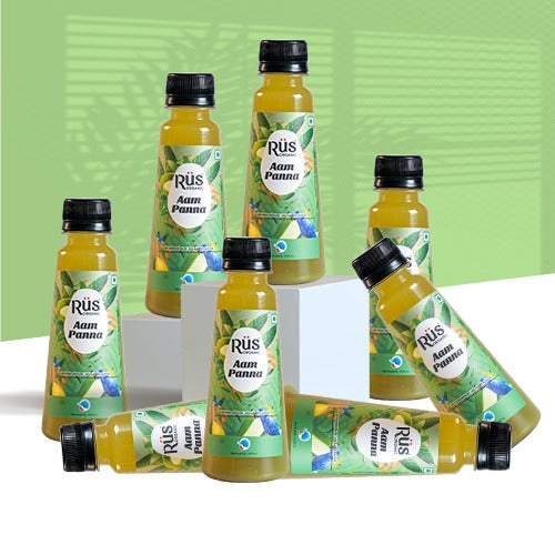 Rus Organic Aam Panna cold-pressed juice is angled on a bed of light-colored sand with scattered seashells and small stones. The bottle has a black cap and a green and light blue label featuring illustrations of green mangoes and butterflies, along with the "Rūs organic aam panna" branding. The background shows a blue sky with wispy white clouds and blurry green palm fronds in the corners.A bottle of Rus Organic Aam Panna juice, emphasizing "no added sugar, no concentrate, no added flavor" on its label. It